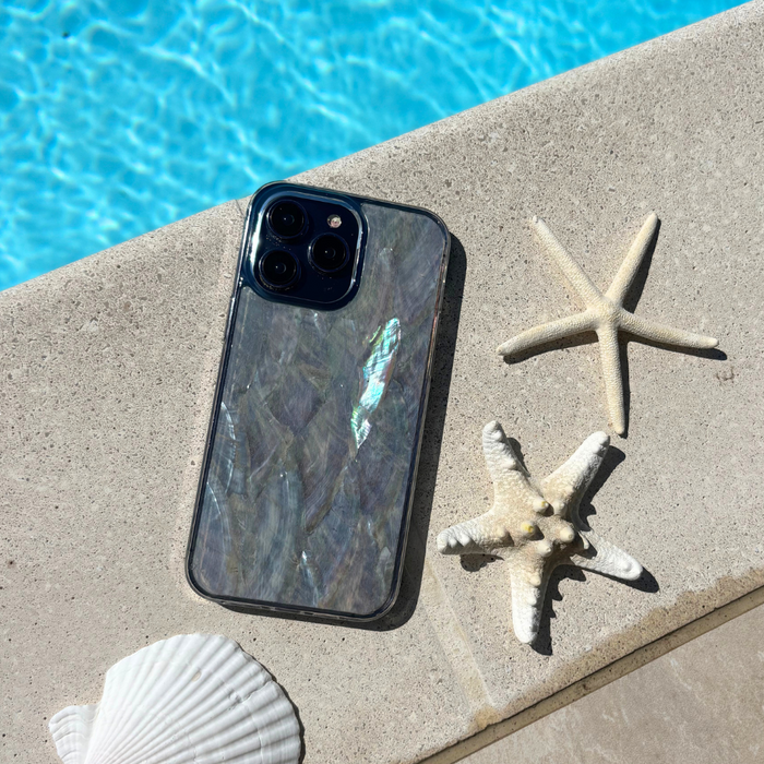 White natural shell phone case with shimmery finish next to seashells and starfish on stone by a pool.