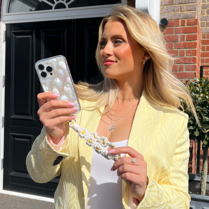 A woman with long blonde hair is standing outside, holding up her phone encased in a Real Natural Shell Phone Case - White by Coconut Lane as if taking a photo or video. She is wearing a pale yellow blazer, a white top, and a necklace. Behind her, there is a black door with white trim and a section of brick wall.