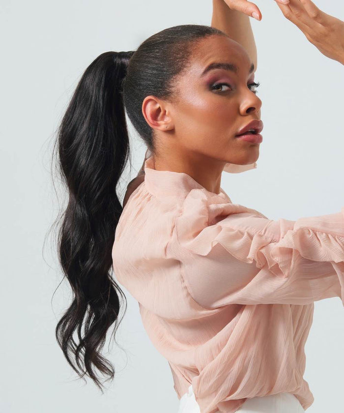 A woman with long, wavy dark hair in a high ponytail using clip-in extensions poses confidently in a sheer pink blouse, one arm raised.