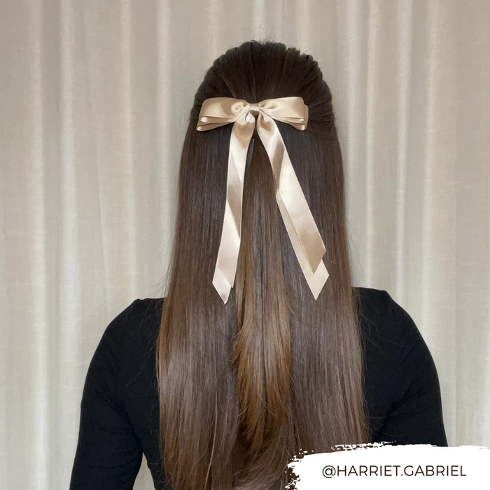 Coconut Lane Caramel Nude ribbon bow hair clip in long, straight brown hair, set against beige curtains.