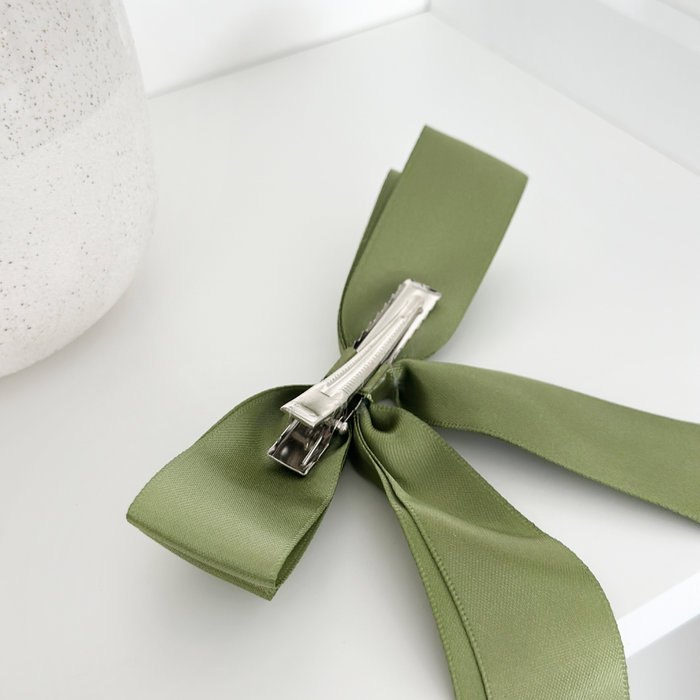 Khaki ribbon bow hair clip with silver barrette next to a textured vase on a white surface.