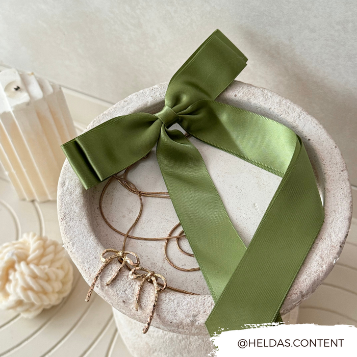 Khaki ribbon bow hair clip with gold earrings and necklace on a stone tray, candle and yarn in the background.
