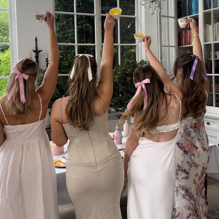 Four women with backs to the camera wear purple Coconut Lane ribbon bow hair clips and hold teacups.