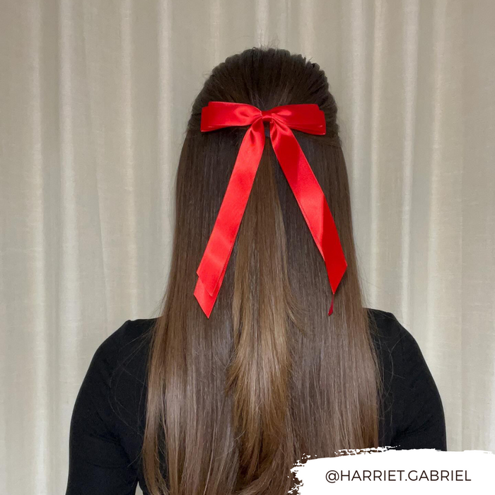 Long brown hair styled half-up with a rose red Coconut Lane ribbon bow clip, shown by beige curtains.