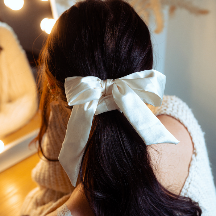 Woman with long dark hair wears an almond cream satin bow scrunchie, seated by a mirror lit with warm lights.