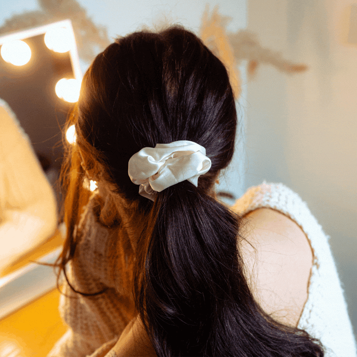 Woman with long dark hair in a ponytail, wearing an Almond Cream satin bow scrunchie by a lit vanity mirror.