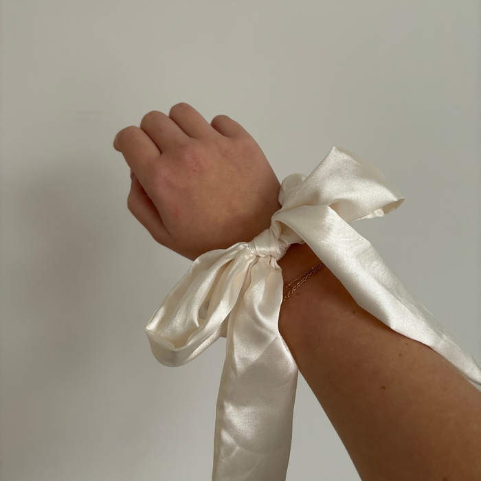 Hand wearing a Coconut Lane satin bow scrunchie in almond cream, shown against a plain background.