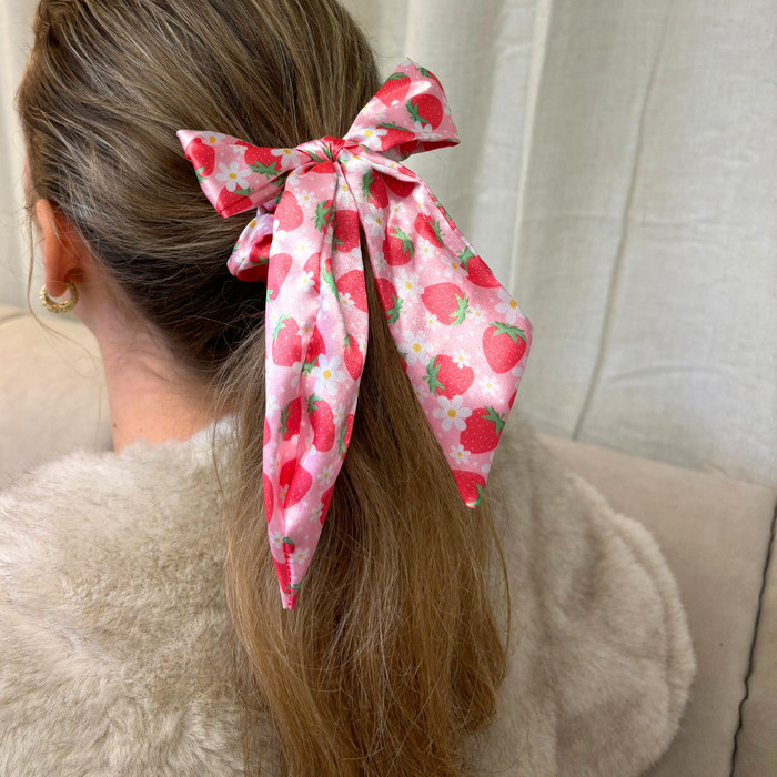 Woman with light brown hair wearing a pink satin bow scrunchie with strawberry print in her ponytail.