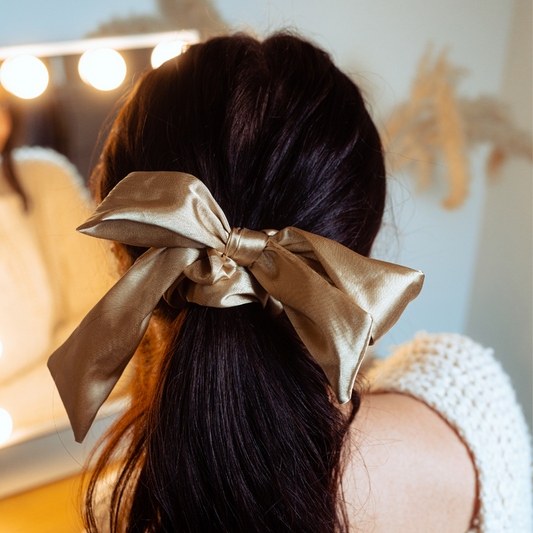 Woman with long dark hair wears a caramel nude satin bow scrunchie, sitting in front of a lighted mirror.