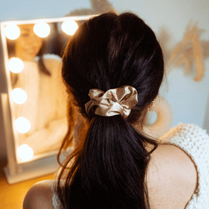 Woman with dark hair in a ponytail wears a caramel nude satin bow scrunchie, standing before a lighted mirror.