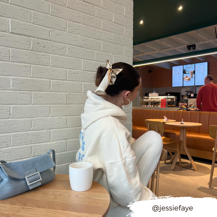 Woman in white hoodie at cafe table, wearing a satin bow scrunchie in caramel nude, holding a coffee cup.