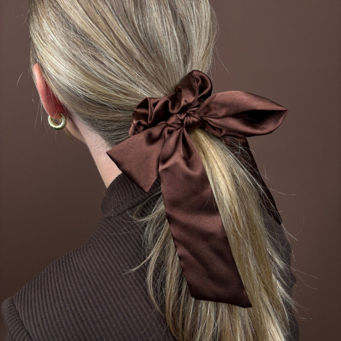 Blonde woman with hair in a ponytail wearing a chocolate brown satin bow scrunchie against a brown background.