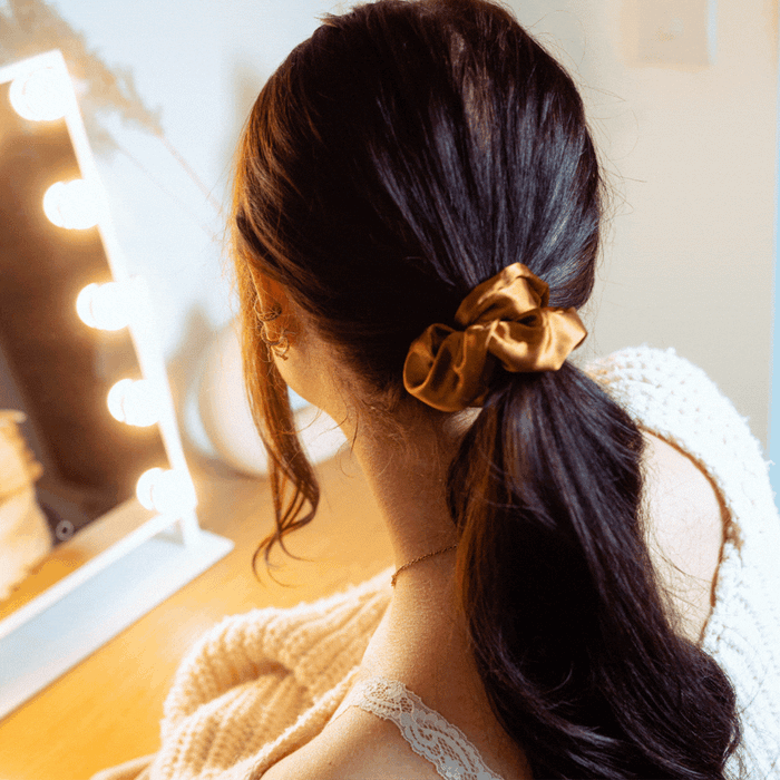 Woman with long dark hair in a ponytail wearing a chocolate brown satin bow scrunchie, beside a lighted vanity.