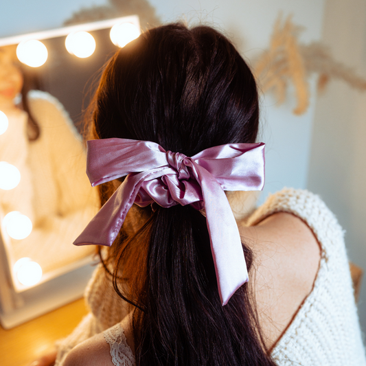 Woman with long dark hair wears a Peony Pink satin bow scrunchie, seated at a vanity in a cream sweater.