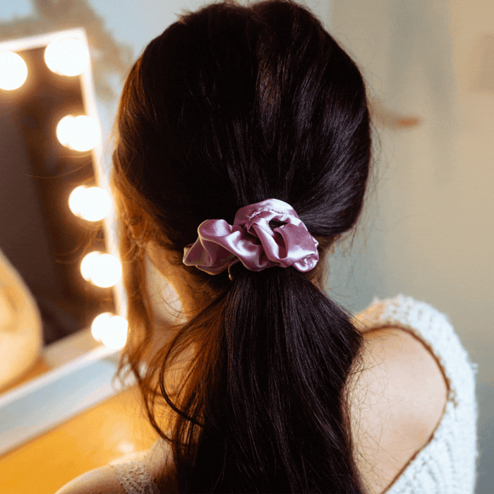 Woman with dark hair in a low ponytail styled with a pink satin bow scrunchie sits at a lit vanity.
