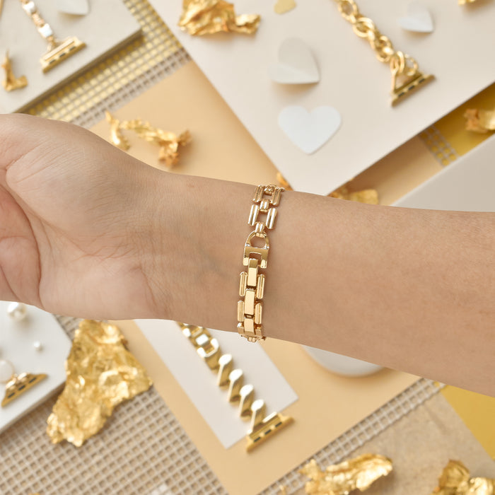A close-up of a person wearing the Square Gold Bracelet Apple Watch Strap by Coconut Lane on their wrist. The background features a yellow and beige theme with gold foil accents, small hearts, and jewelry pieces like necklaces and rings displayed on white and beige surfaces.