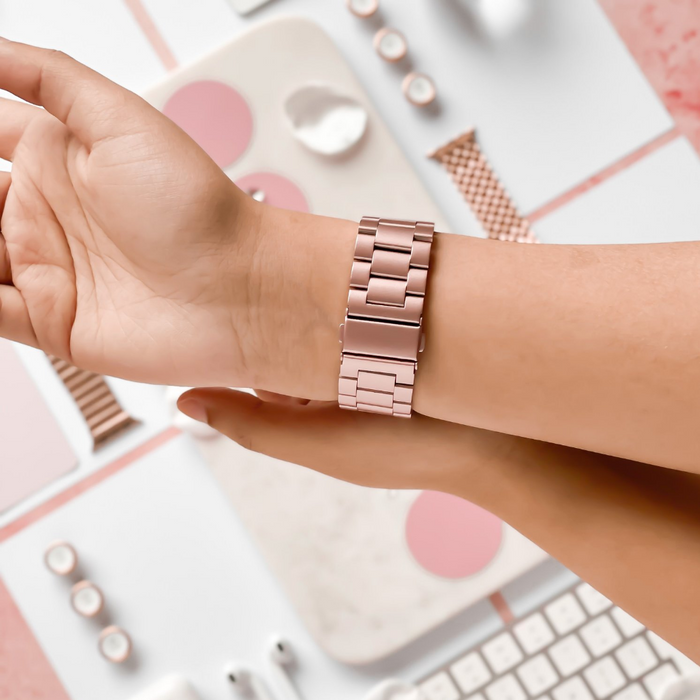 A wrist adorned with a blush-colored stainless steel Apple Watch strap by Coconut Lane is shown in close-up against a backdrop of light pink and white office supplies, including a keyboard, mouse, and decorative items. The adjustable strap features a sleek, modern design with interlocking links.