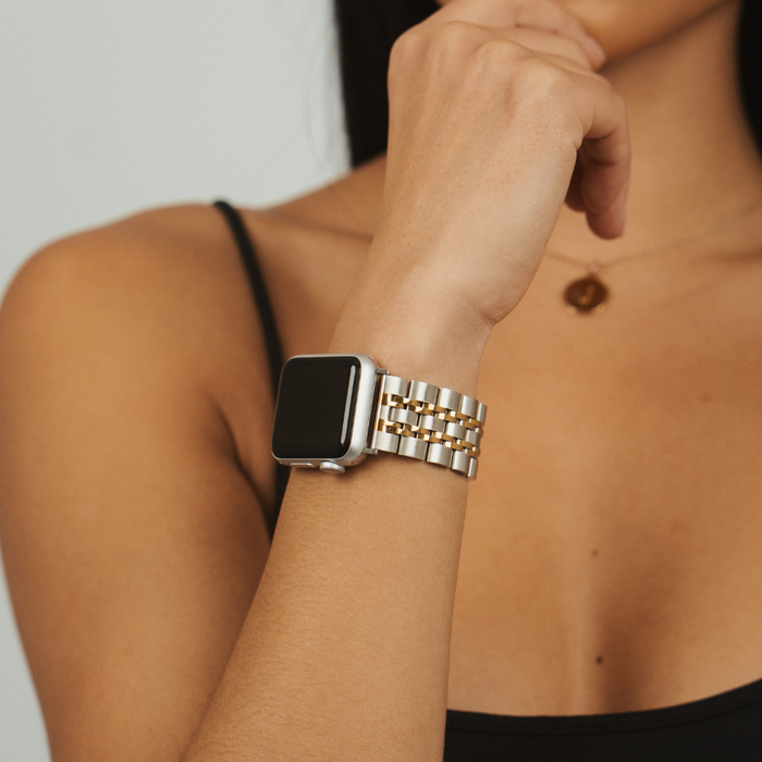 Woman in a black tank top wears a silver and gold stainless steel Apple Watch strap, hand raised near her face.