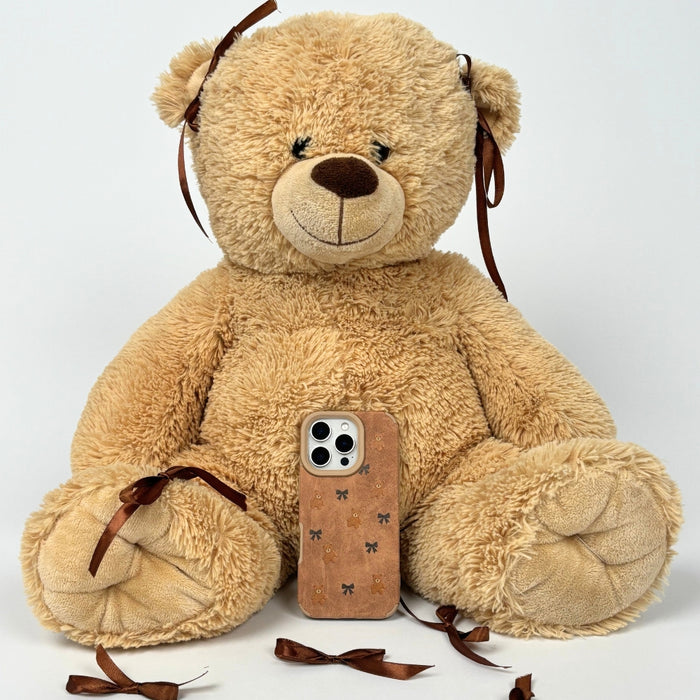 Coconut Lane Bears & Bows suedette phone case with bear and bow pattern, shown with a plush teddy bear in the background.