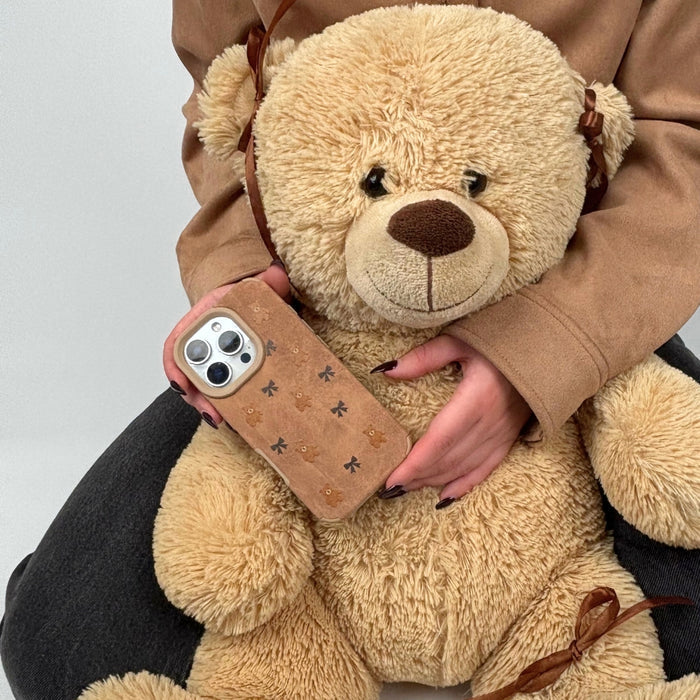 Person sitting on the floor holding a beige teddy bear and a Coconut Lane suedette phone case with bears and bows pattern.