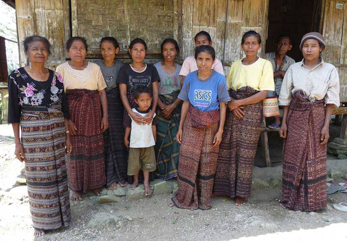 Group of women and a child standing in front of a wooden workshop where symplocos mordant is made by hand.