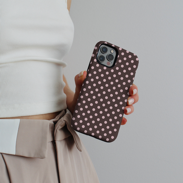 Hand holding a brown polka dot phone case; person wears a white top and beige pants.