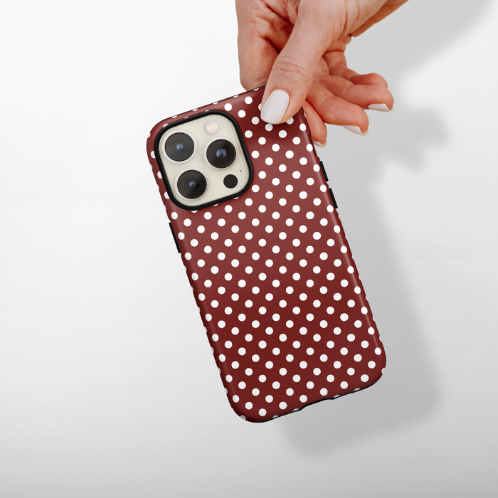Hand holding a burgundy polka dot MagSafe phone case by Coconut Lane against a white background.