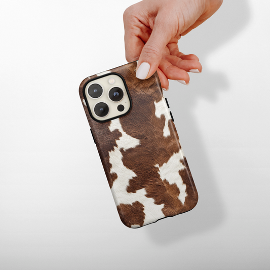 Hand holding a Cocoa Cowgirl cow print MagSafe® phone case by Coconut Lane, shown against a light background.
