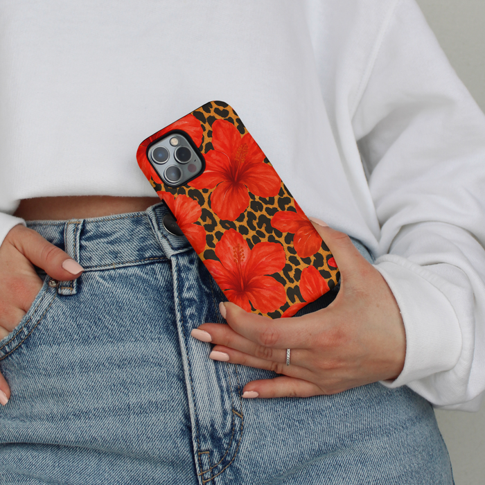 Person in jeans and a white sweatshirt holding a tough phone case with bold red leopard hibiscus print.