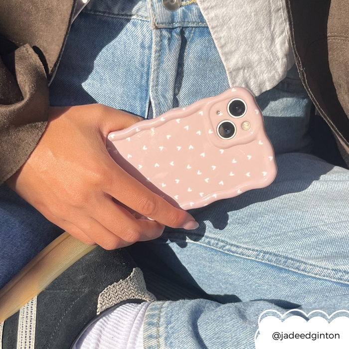 Hand holding a Coconut Lane phone case with wavy chai-colored hearts; person wears jeans, a jacket, and black sneakers.