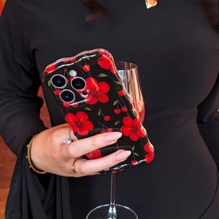 Hand with rings and necklace holds a wine glass and a wavy pink phone case with cherry hibiscus pattern.