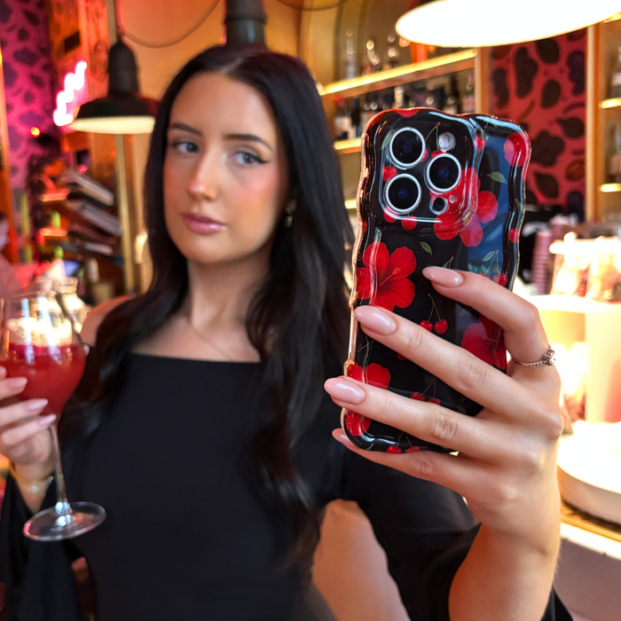 Woman with long dark hair takes a selfie in a cafe, holding a phone in a cherry and white wavy patterned case.