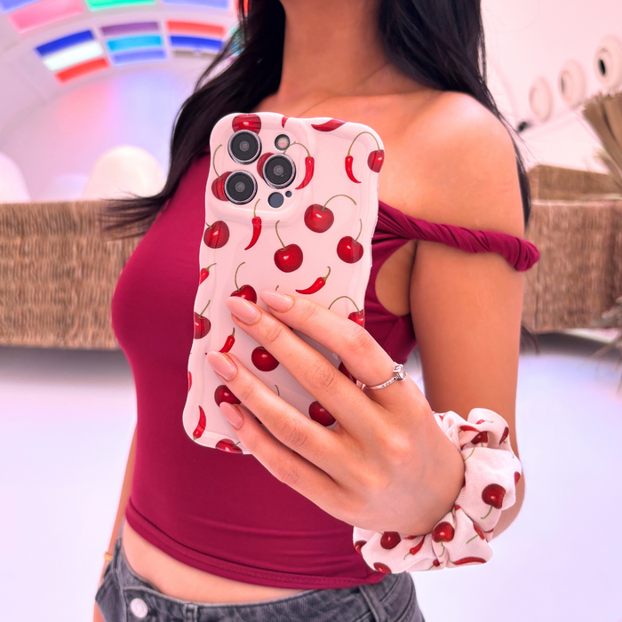 Woman in maroon top holds red wavy Coconut Lane phone case with cherry pattern, matching her hair scrunchie, in mirror selfie.