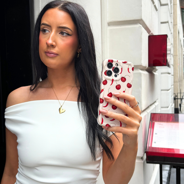 Woman wearing white off-shoulder top takes selfie holding red wavy phone case with gold necklace visible.