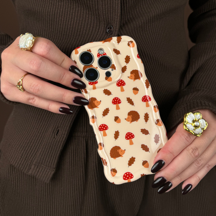 Hand with dark nails and rings holding a wavy phone case featuring hedgehogs, mushrooms, and leafy forest patterns.