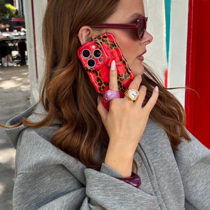 Woman in a gray hoodie holding a wavy Coconut Lane phone case with a leopard and hibiscus pattern, wearing rings and sunglasses.