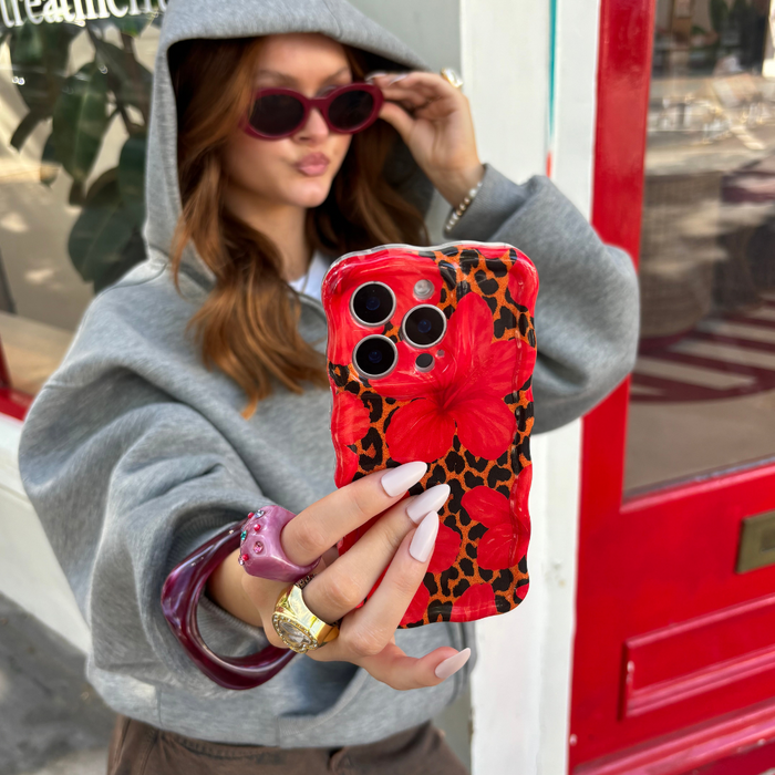 Woman in gray hoodie holds a Coconut Lane phone case with a wavy leopard and hibiscus pattern, wearing sunglasses.