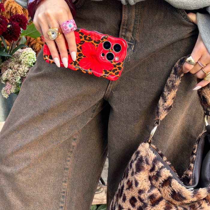 Person in brown jeans holds Coconut Lane leopard hibiscus wavy phone case and a leopard-print bag, wearing colorful rings.