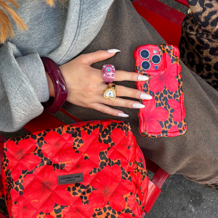 Hand with chunky rings holds a wavy leopard floral phone case beside a matching bag on a red chair.