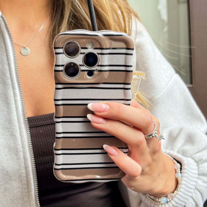 Hand holding Coconut Lane phone case with retro wavy stripes, manicured nails, rings, and gray hoodie visible.