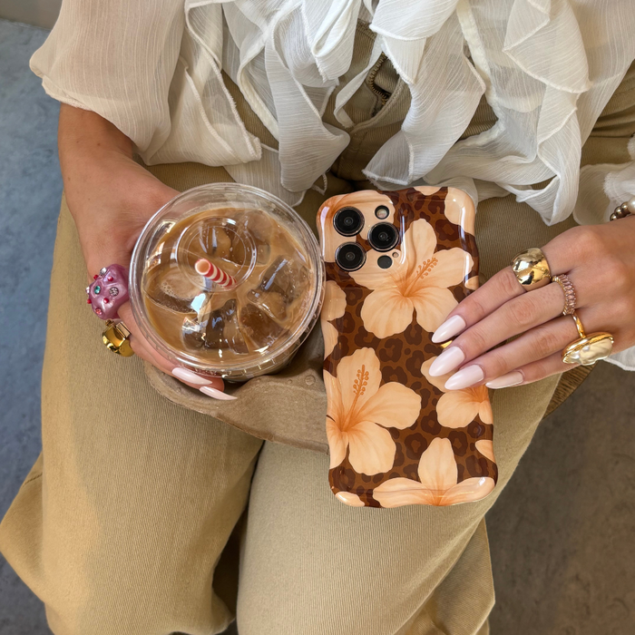 Hand holding an iced coffee and a phone with a wavy, floral Coconut Lane Safari Bloom case. Gold rings and beige pants visible.
